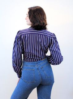 "This is a marvelous 1970s vintage blouse that has a lot going for it! The fabric is a silken poly with a very pleasant soft matte hand, like a charmeuse. The pattern is a classic vertical bar stripe in navy blue with taupe and a bright red pinstripe. It has shirring at the front shoulder yokes, puffed shoulders with small shoulder pads, long sleeves with notched single button cuffs, an open V neckline with a notched collar, and it looks great tied in front or worn with the straight hem down. Ou Fitted Button-up Tops With Vertical Stripes, Fitted Vertical Stripe Button-up Tops, Vintage Collared Top With Vertical Stripes, Vintage Collared Tops With Vertical Stripes, Vintage Puff Sleeve Blouse With Buttons, Fitted Button-up Blouse With Vertical Stripes, Fitted Vertical Stripes Button-up Blouse, Vintage Striped Fitted Tops, Vintage Fitted Striped Tops