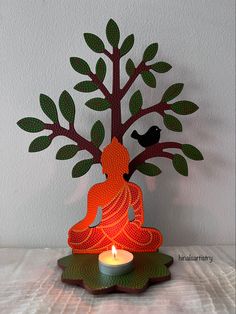 a lit candle is sitting in front of a paper cut out buddha statue with a tree on top