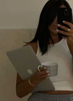 a woman is holding a laptop and looking at her cell phone while she holds a coffee mug