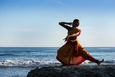 a woman standing on top of a rock next to the ocean wearing a yellow and red dress