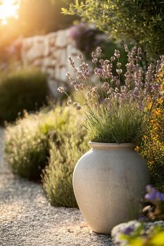 a large white vase sitting on top of a gravel road next to bushes and flowers