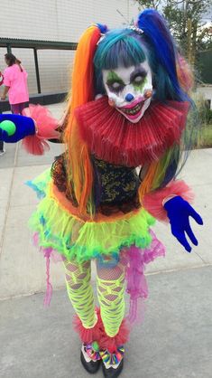 a woman dressed in clown makeup and colorful hair is standing on the sidewalk with her hands behind her back