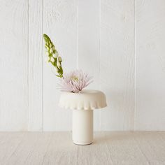 a white vase with a pink flower in it sitting on a table next to a wall