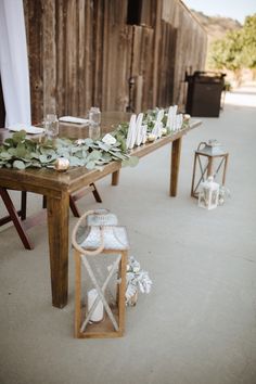 a wooden table topped with white flowers and greenery next to a barn wall filled with candles