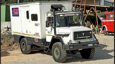 a white truck parked in front of a building with other trucks behind it on a dirt road