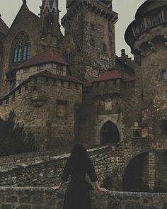 a woman is standing in front of an old castle with stone walls and towers, looking down at the ground