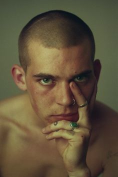 a shirtless man with piercings on his nose and hand under his chin looking at the camera