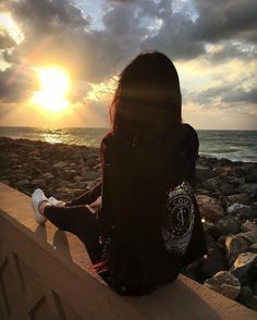 a woman sitting on the edge of a wall looking out at the ocean while the sun is setting