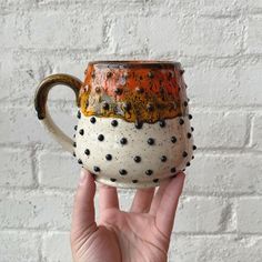 a hand holding up a ceramic mug with holes on the outside and inside, in front of a white brick wall