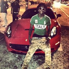 a man sitting on the hood of a red sports car in front of some people