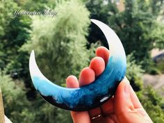 a hand holding a fake crescent moon in front of some trees and bushes on a sunny day