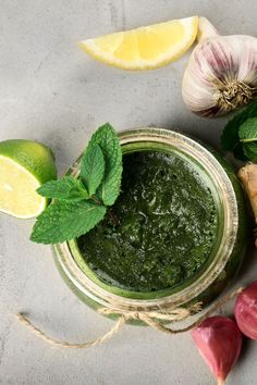 a jar filled with green pesto next to garlic and lemon wedges on top of a table