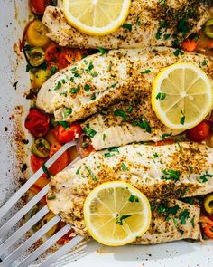 two fish with lemons, tomatoes and peppers in a white dish next to a fork