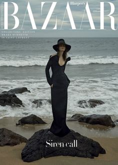 a woman standing on top of a rock next to the ocean wearing a black dress and hat