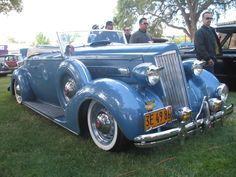 two men standing next to an old blue car in the grass at a car show