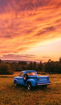 an old blue pickup truck parked in a field with the sun setting behind it and clouds