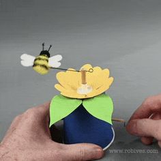 a hand holding a piece of paper with a flower on it and a bee flying over it