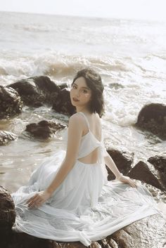 a woman sitting on top of a rock next to the ocean wearing a white dress