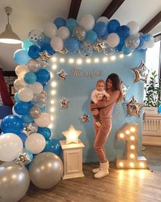 a woman holding a baby standing in front of a backdrop with blue and white balloons