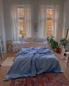 a bed with blue sheets and pillows on top of a wooden floor next to two windows