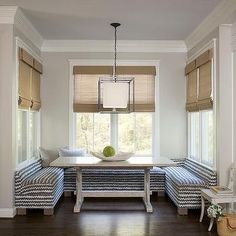 a dining room table and bench in front of two windows with roman shades on them