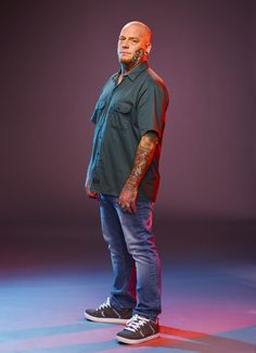 a bald man with tattoos standing in front of a purple and blue background wearing jeans