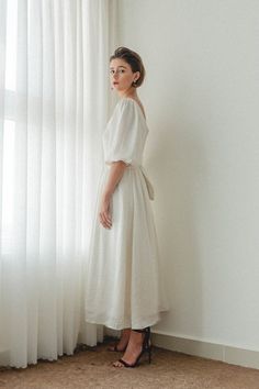 a woman standing in front of a window wearing a white dress and black heels with her hands on her hips