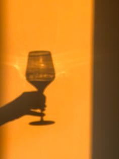 the shadow of a hand holding a wine glass in front of a yellow wall with light coming through it