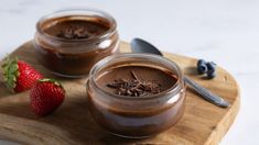 two jars of chocolate pudding on a cutting board with a strawberry and spoon next to it