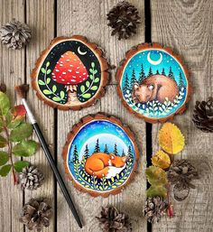 three painted wood slices with leaves and pine cones around them on a wooden table next to an acorn