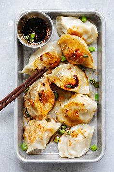 a tray filled with dumplings covered in sauce and green onions next to chopsticks