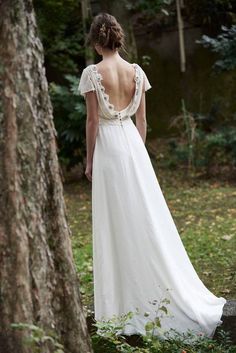 the back of a woman in a white dress standing next to a tree with her hands on her hips