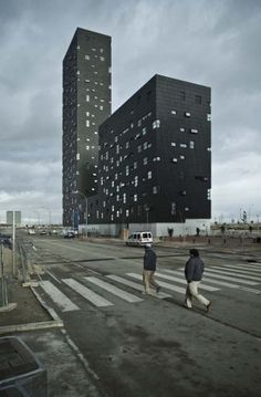 two people walking across an empty parking lot in front of tall buildings with square windows