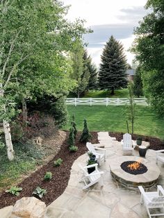 a fire pit surrounded by white chairs and trees in a yard with green grass, rocks and shrubs