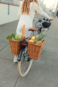 a woman riding on the back of a bike next to a basket filled with fruit