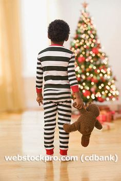 a little boy in striped pajamas holding a teddy bear while standing next to a christmas tree