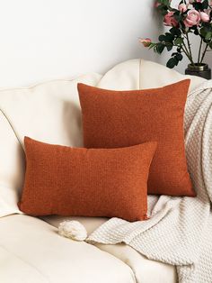 two orange pillows sitting on top of a white couch next to a vase with flowers