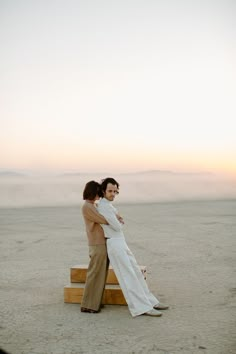 two people standing on top of a wooden bench in the middle of a sandy field