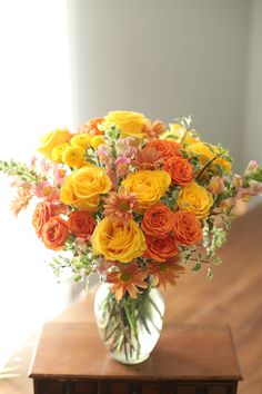 a vase filled with orange and yellow flowers on top of a wooden table next to a window