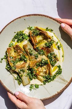 two hands holding a white bowl filled with artichokes and sauce on top