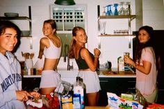 four young women standing in a kitchen next to a counter with food on it and bottles