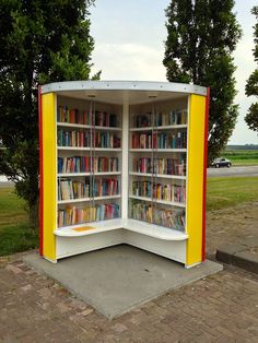 a yellow and white book case sitting in the middle of a park with lots of books on it