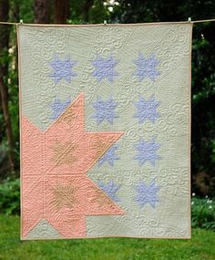 a pink and blue star quilt hanging from a clothes line in the grass with trees in the background