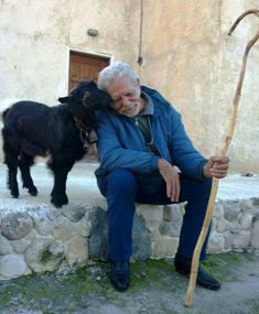 an old man sitting on a stone bench next to a small black goat and holding a stick