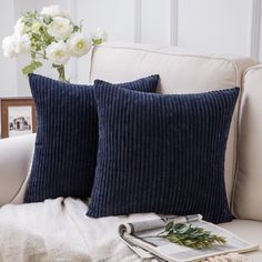 two blue pillows on a white couch with flowers in a vase next to the pillow