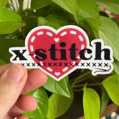 someone holding up a sticker with the word x stitch on it in front of some plants