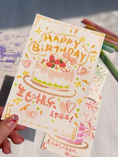 a person holding up a birthday card in front of some colored pencils and paper