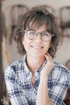 a woman with glasses is smiling at the camera while wearing a plaid shirt and holding her hand to her chin