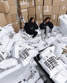 two women sitting on cardboard boxes surrounded by white bags with black words that read the same