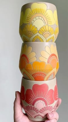 a hand holding three different colored vases in each other's hands, one with yellow and red flowers on it